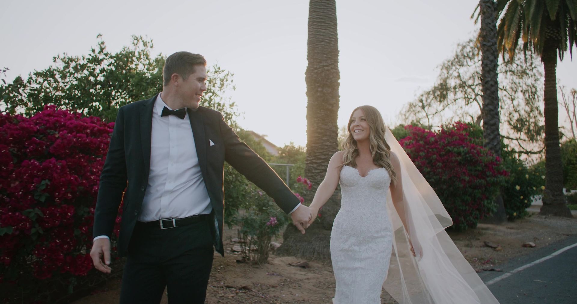 Shane and Rosie walking hand in hand past a wall of bougainvillea and palms.