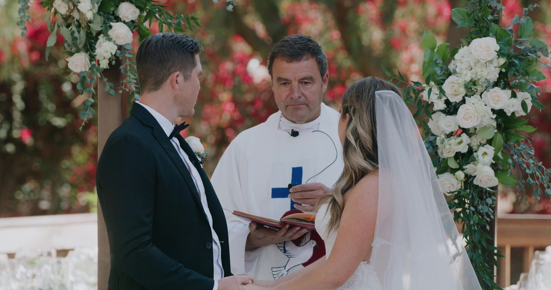 The ceremony at the floral arch, Shane and Rosie facing each other with the officiant between them.