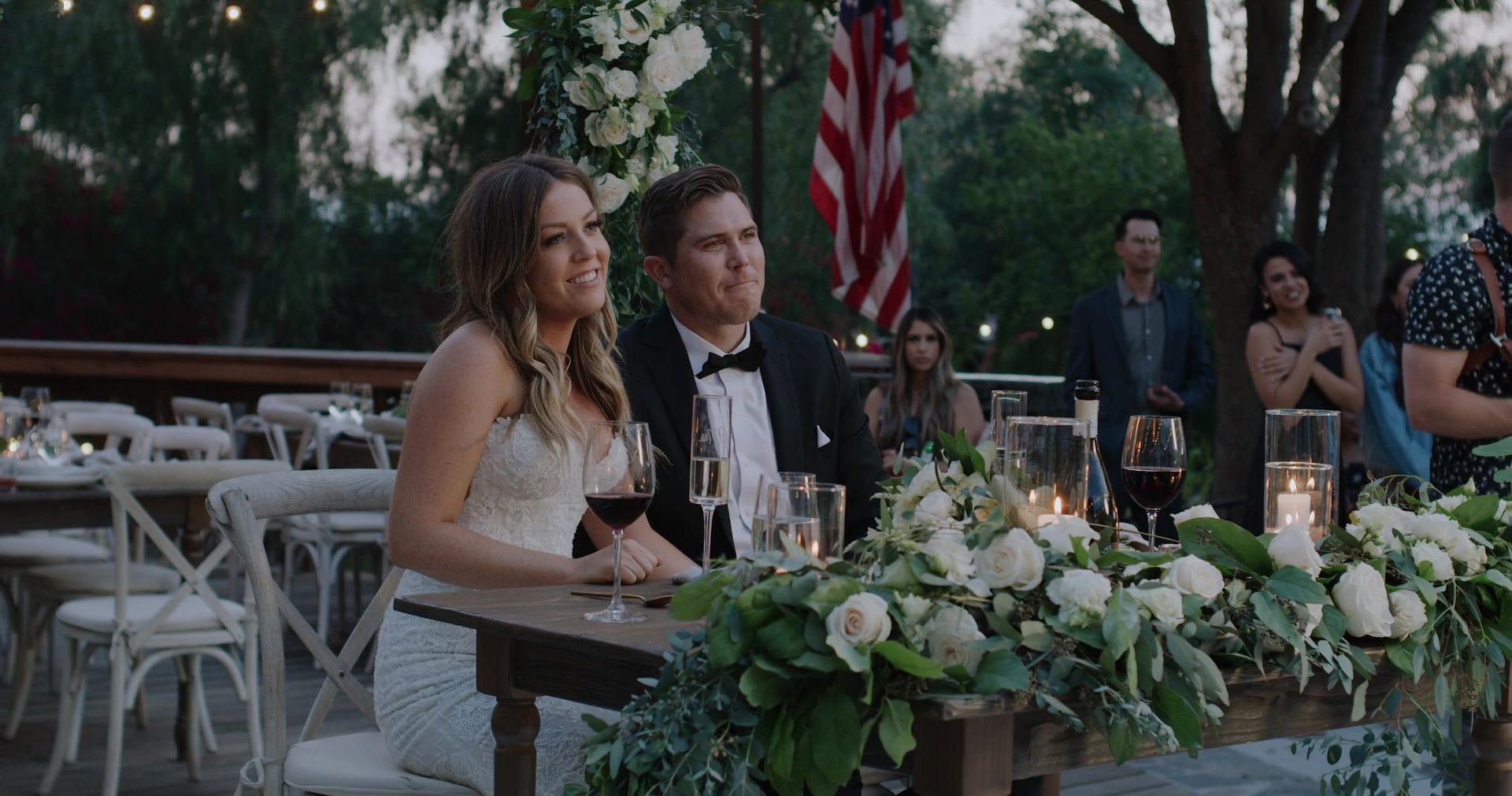 Shane and Rosie seated at their table, a garland of white roses and greenery along its front and an American flag hanging behind.