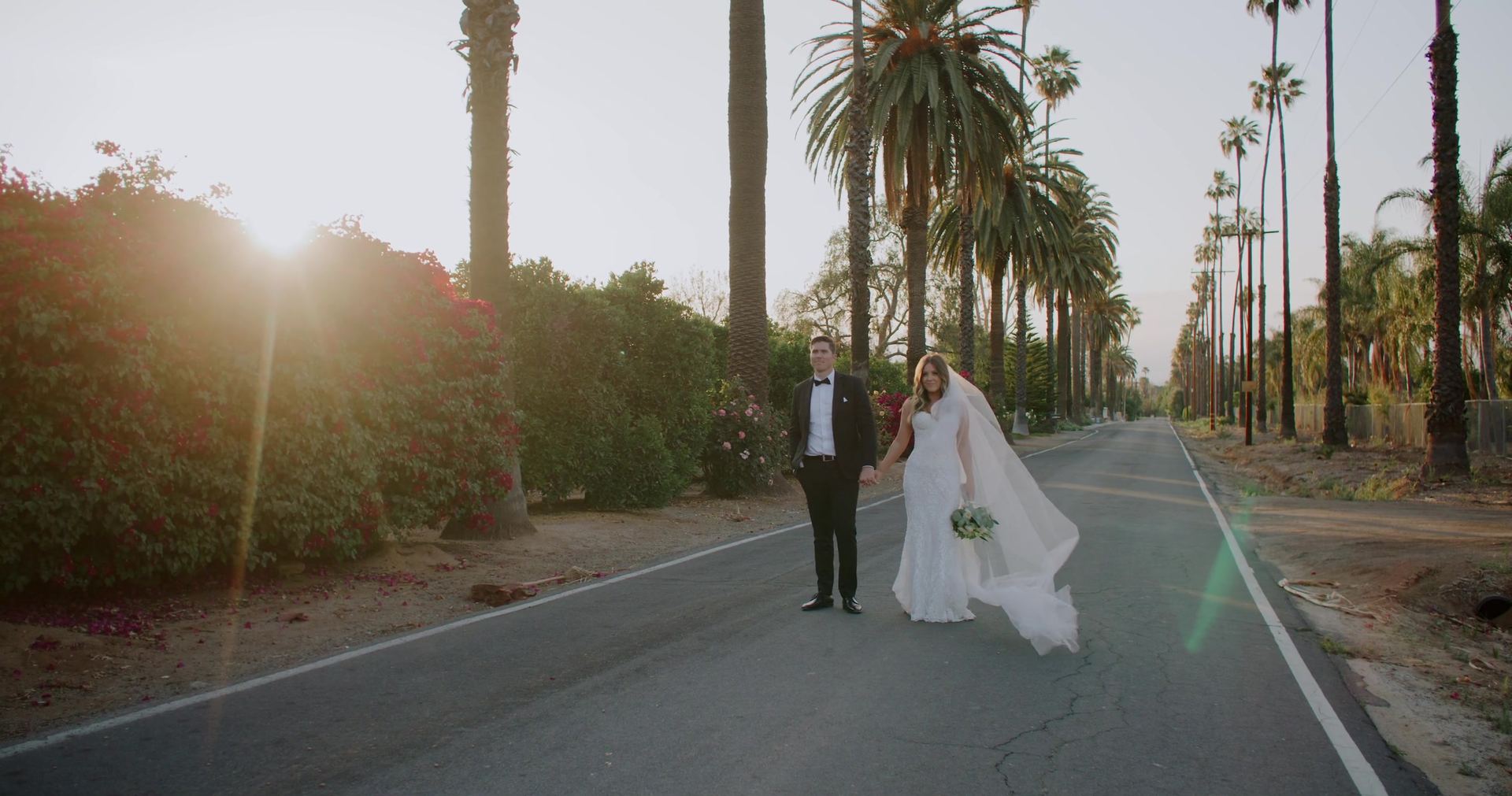 Shane and Rosie hand in hand on a palm-lined road, her veil lifting, the low sun flaring through bougainvillea at the left.