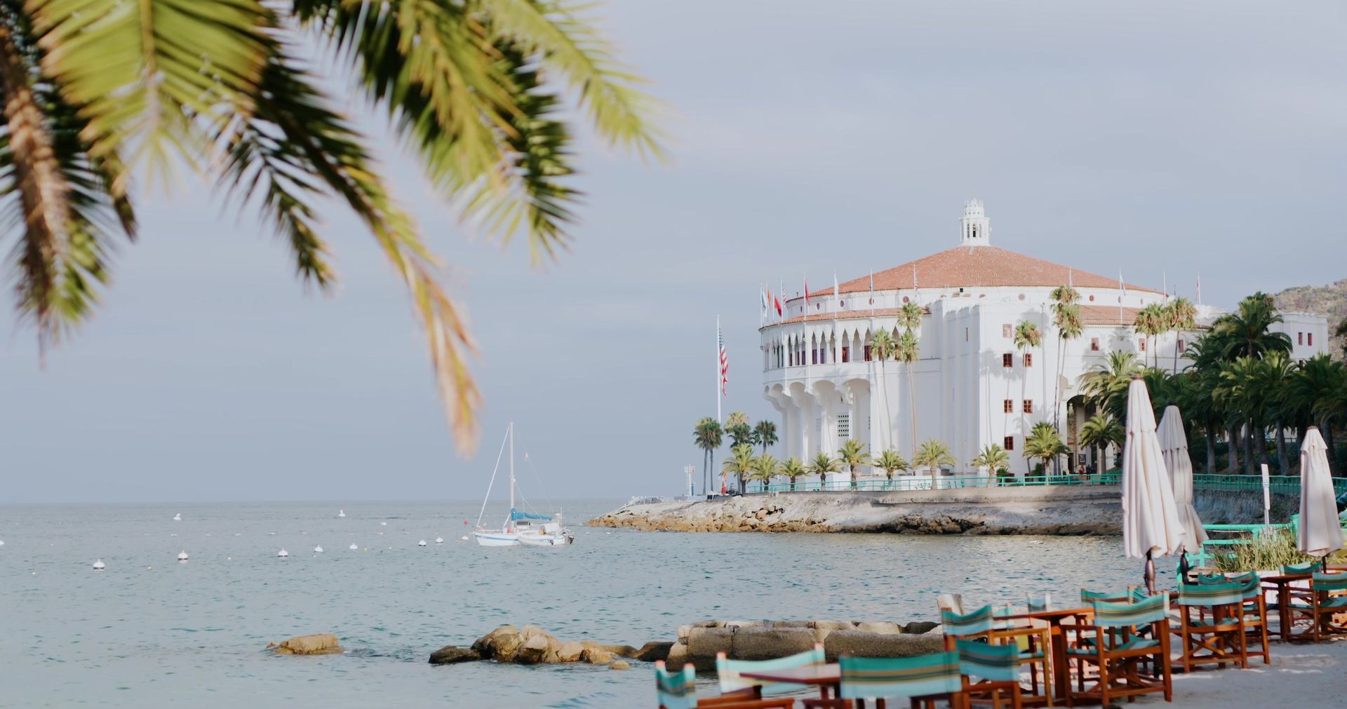 The round white Casino building seen past palm fronds, a sailboat moored in the bay and rows of striped chairs along the shore.