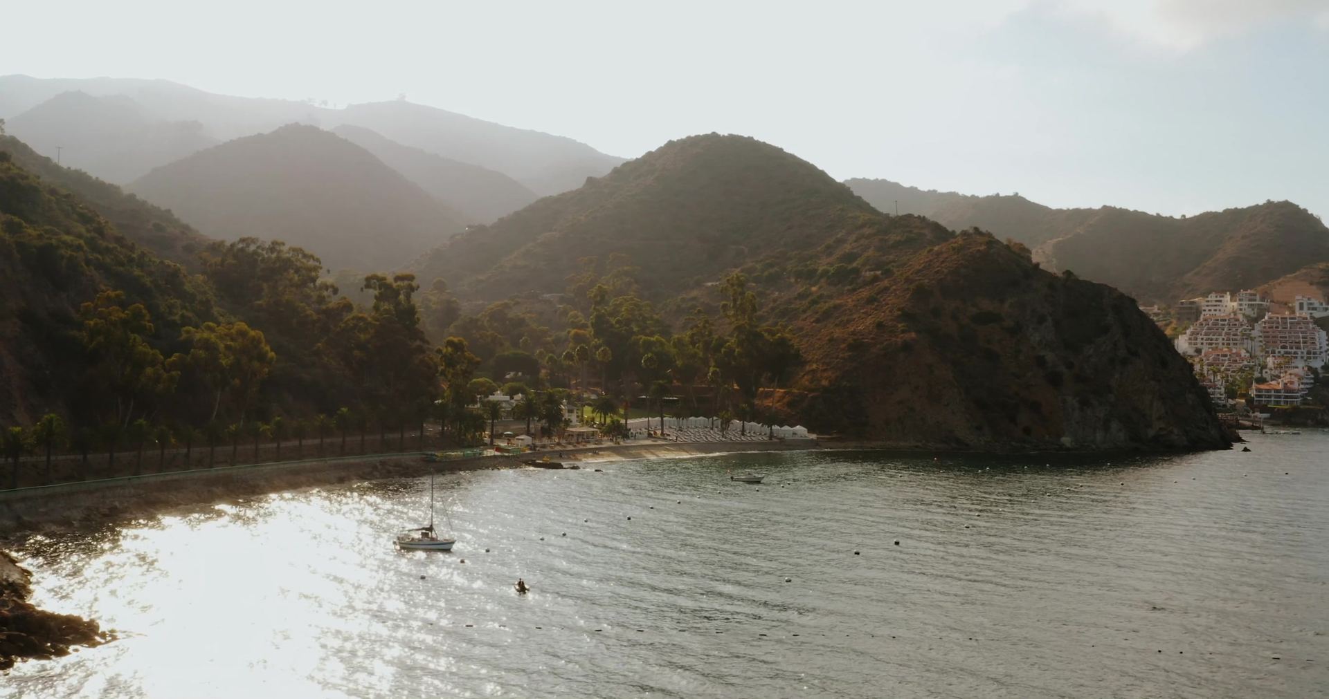 The cove in hazy backlight, a beach lined with white cabanas below the hills, a sailboat moored offshore and white buildings on the far point.