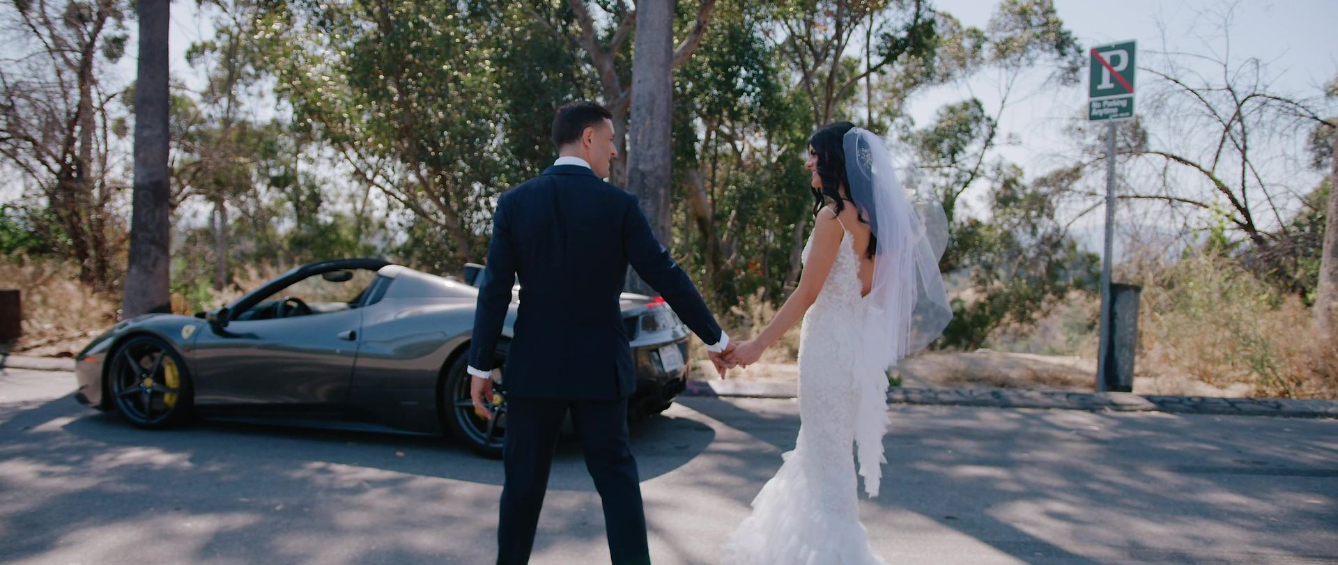 Tyler and Eliza hand in hand beside a grey convertible sports car parked on a road beneath eucalyptus trees.
