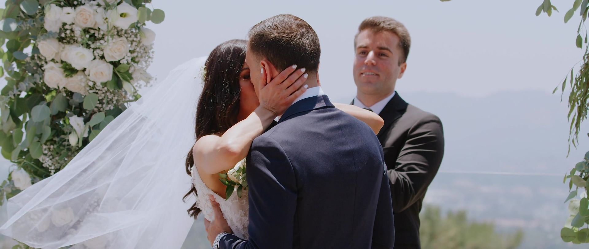 The kiss under the floral arch, the officiant smiling beside them and hazy hills below.