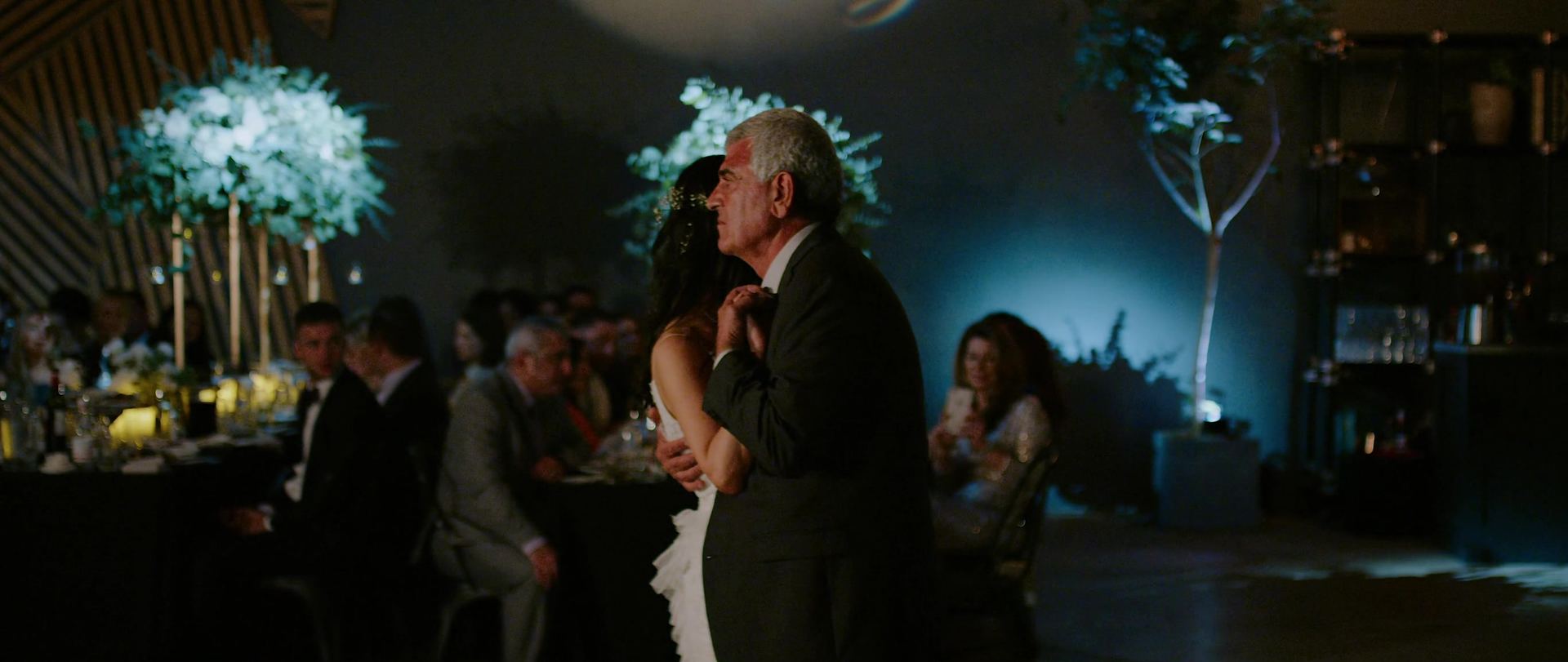 Eliza dancing with a silver-haired man in a grey suit on a dim dance floor, tall uplit greenery arrangements on the tables behind.