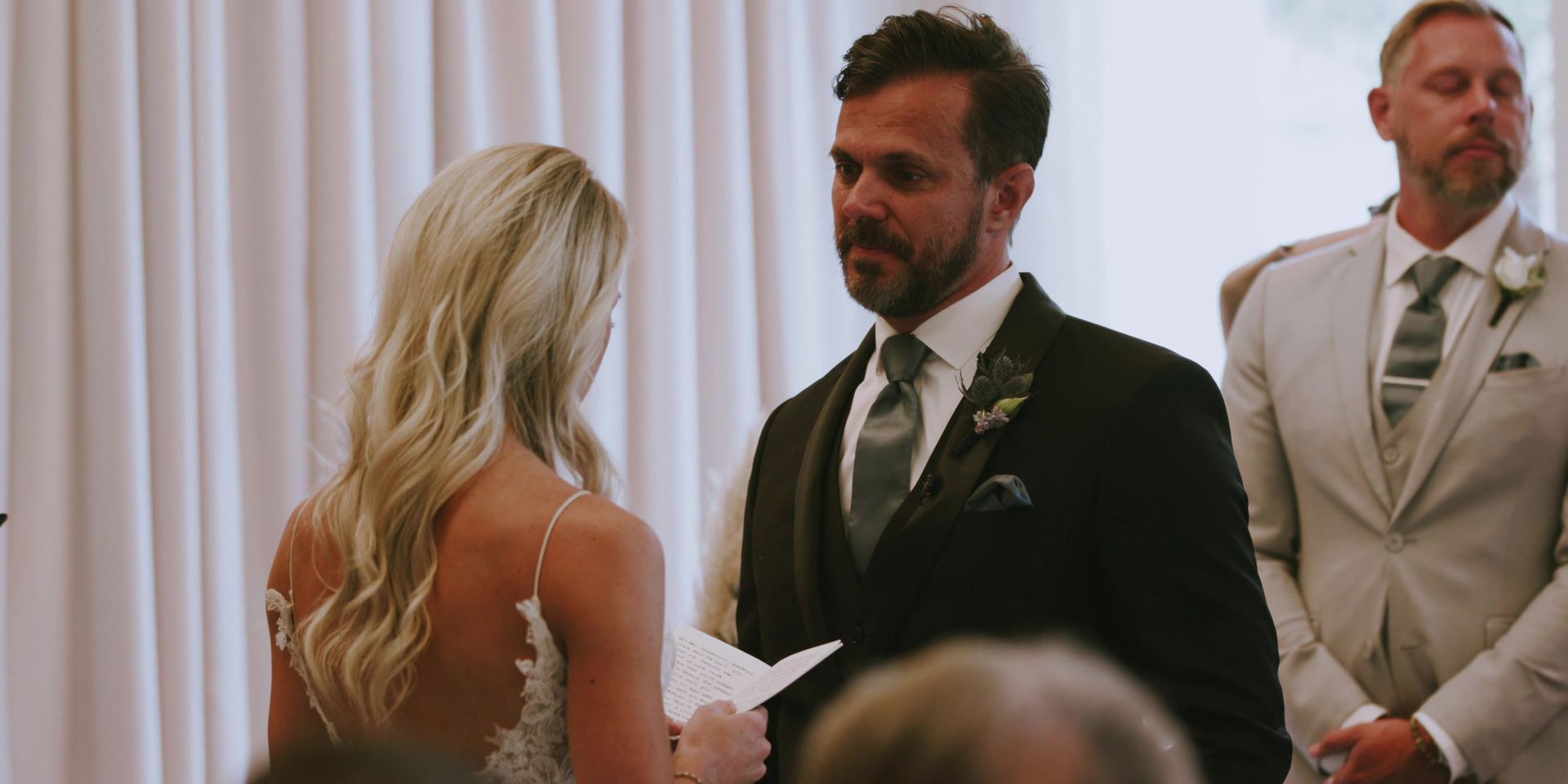 Katherine reading her vows aloud from a card, Tony listening, a groomsman in a light grey suit standing behind him.