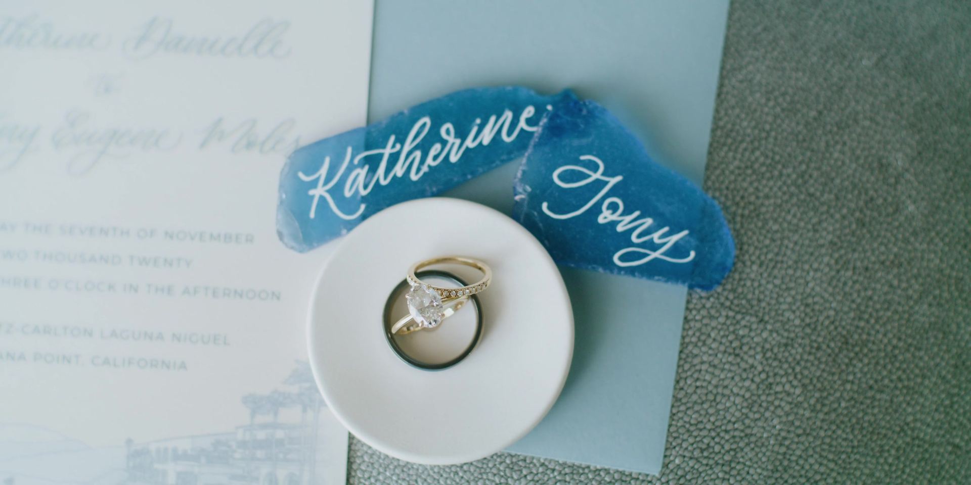 The invitation and rings, two pieces of blue glass calligraphed Katherine and Tony beside a white dish holding an engagement ring and two bands.