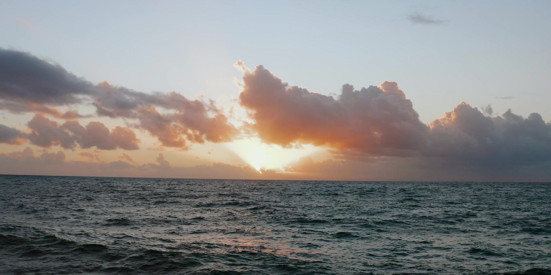 The sun low behind a bank of clouds over open water, light breaking through above the horizon.