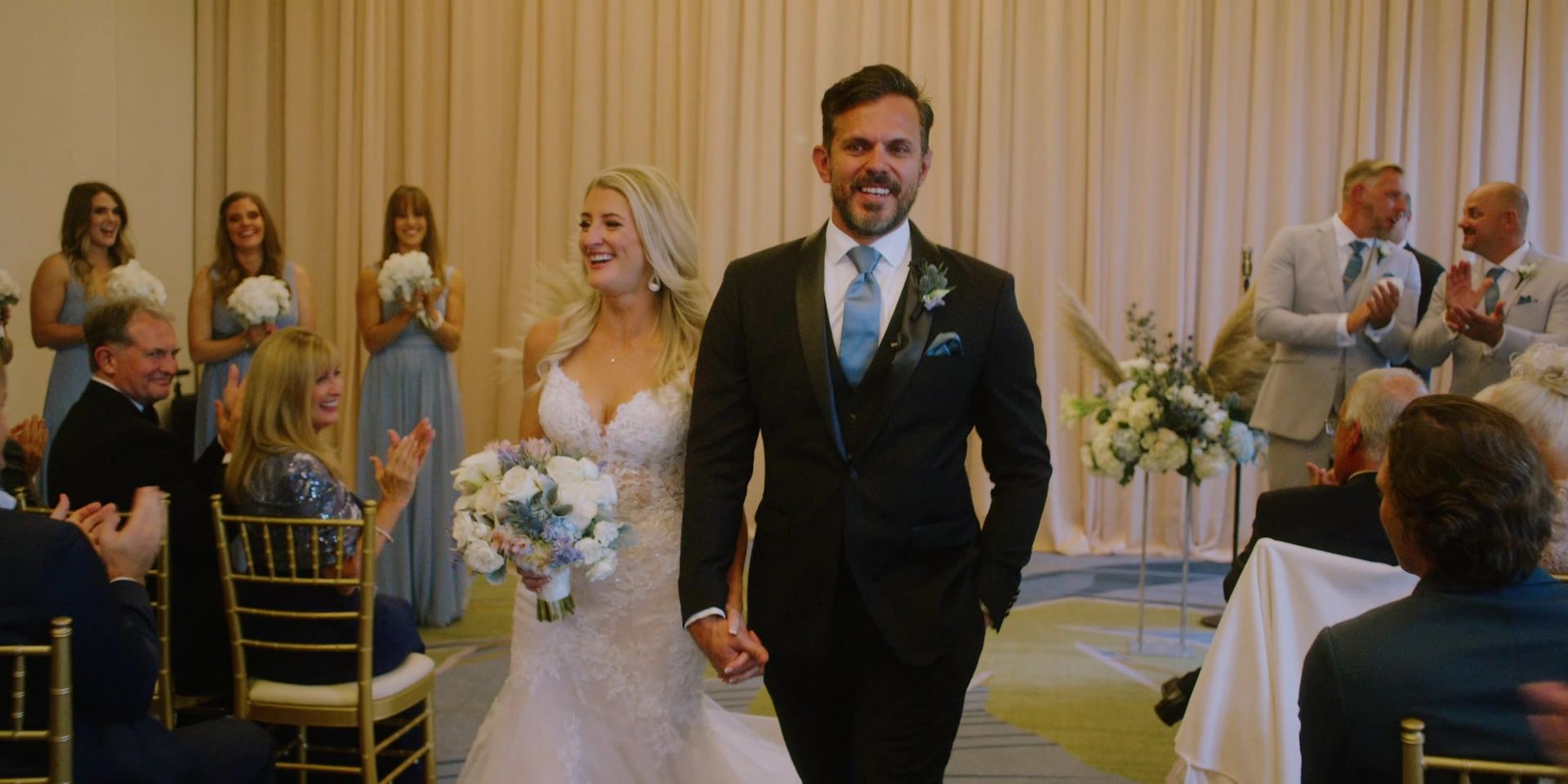 Tony and Katherine walking back up the aisle hand in hand, guests applauding from the chairs on both sides.