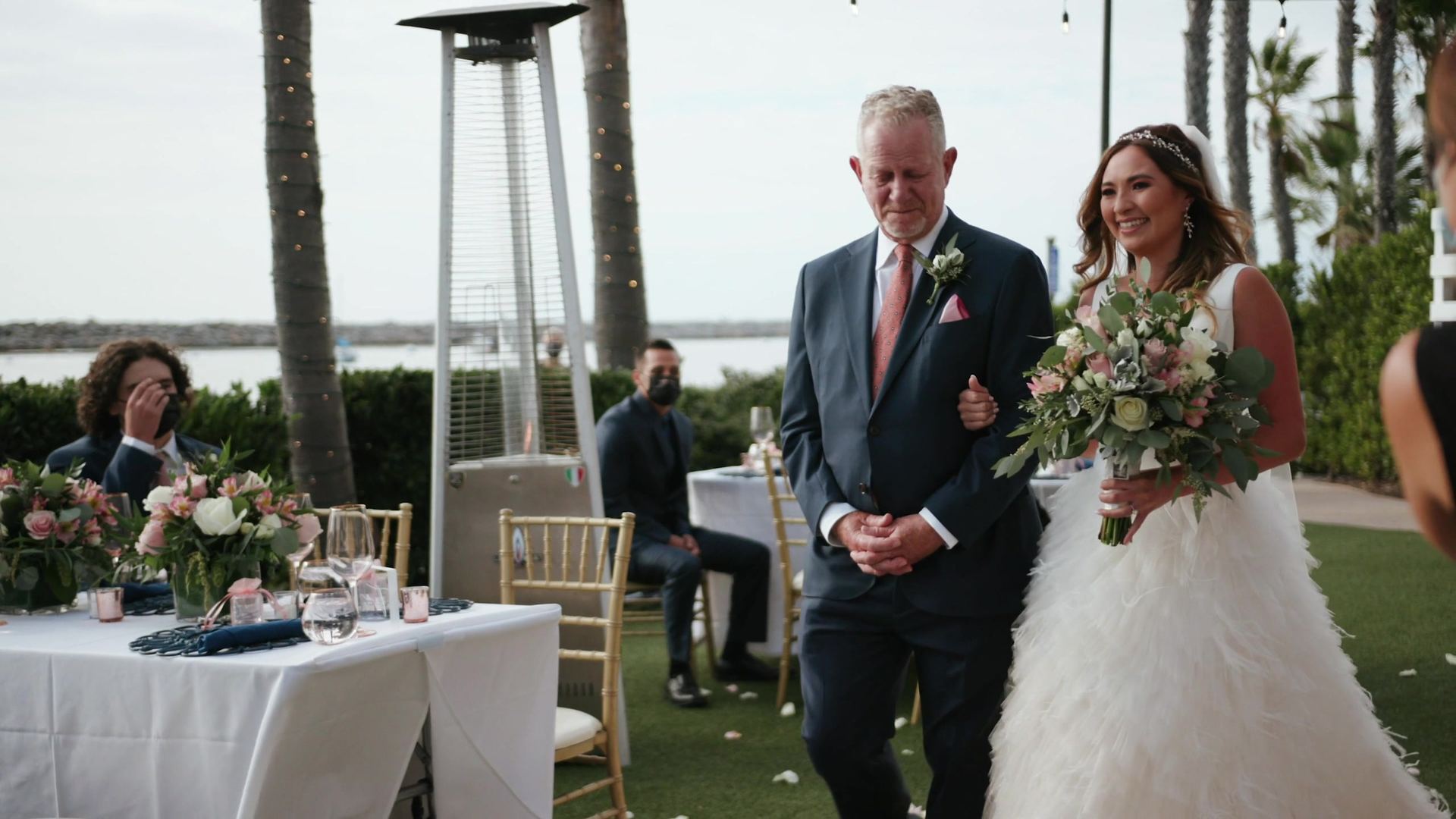 Rachel coming in on the arm of a grey-haired man in a navy suit, past the set tables and a patio heater, the breakwater and water behind.