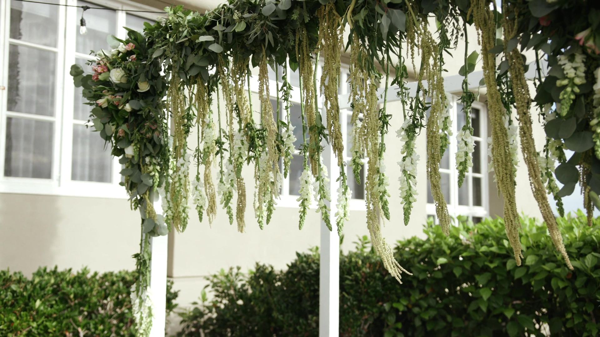 The ceremony arch, hanging amaranthus, white snapdragons and eucalyptus draped from a white frame in front of the hotel's cream wall and paned windows.