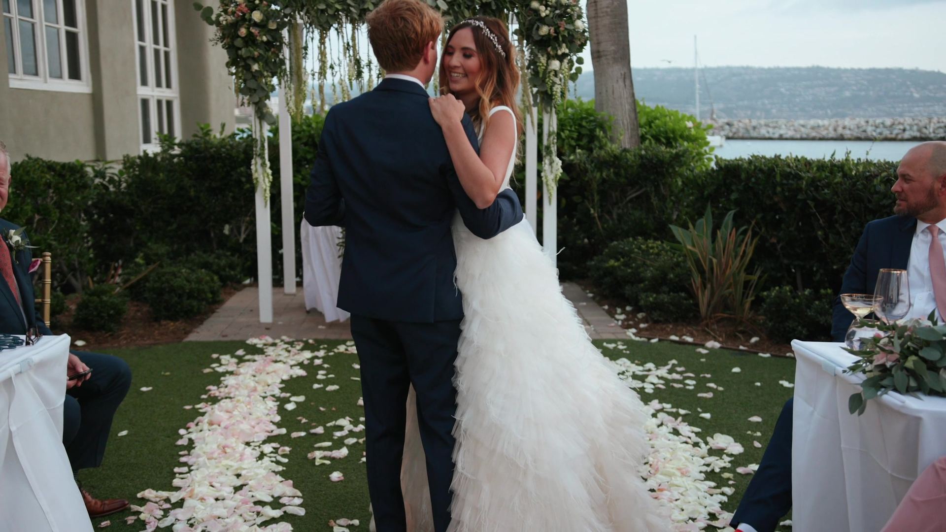 Tyler and Rachel dancing on the petal-strewn ceremony lawn beneath the floral arch, the breakwater and water beyond.