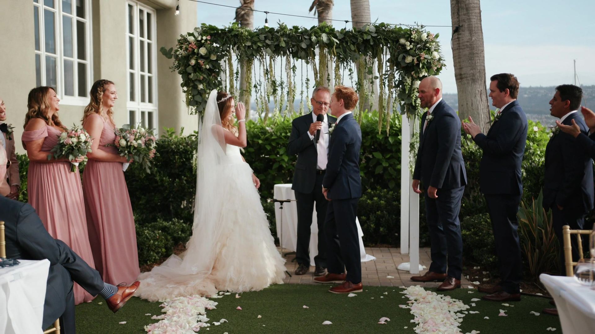 Tyler and Rachel at the altar as the officiant speaks into a microphone, the wedding party on either side and petals lining the aisle.