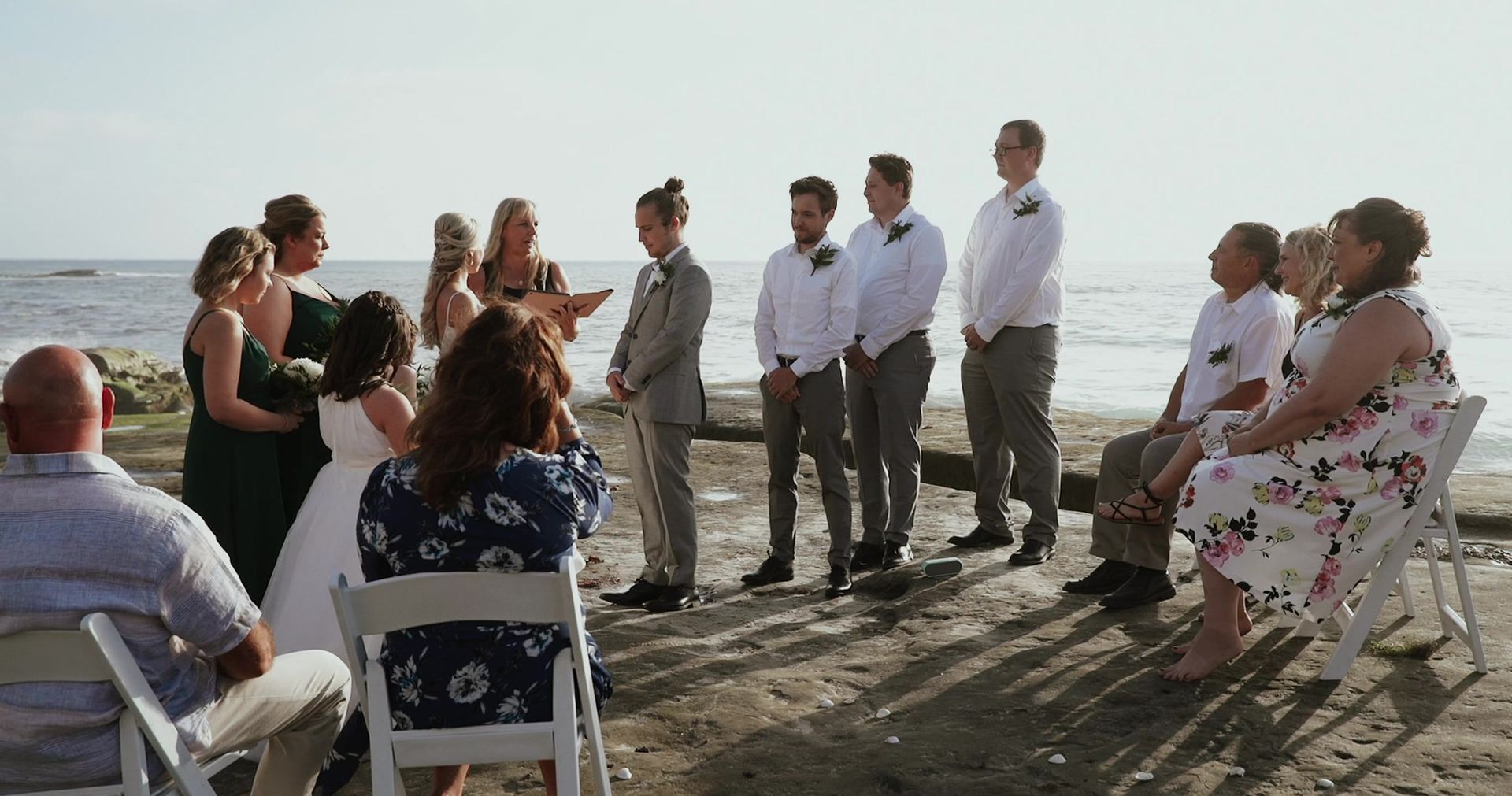The ceremony on the beach, the groomsmen in white shirts and grey trousers beside Mitch, bridesmaids in green, guests seated in white folding chairs with the ocean behind.