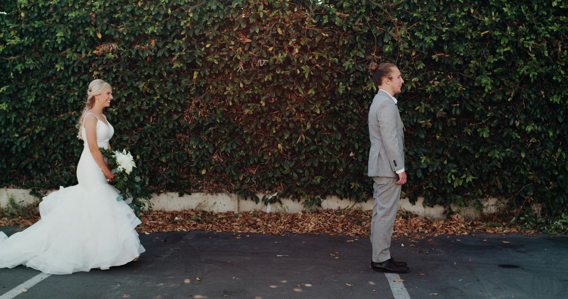 Zoe and Mitch facing each other across an empty parking lot in front of an ivy-covered wall, her bouquet at her side.
