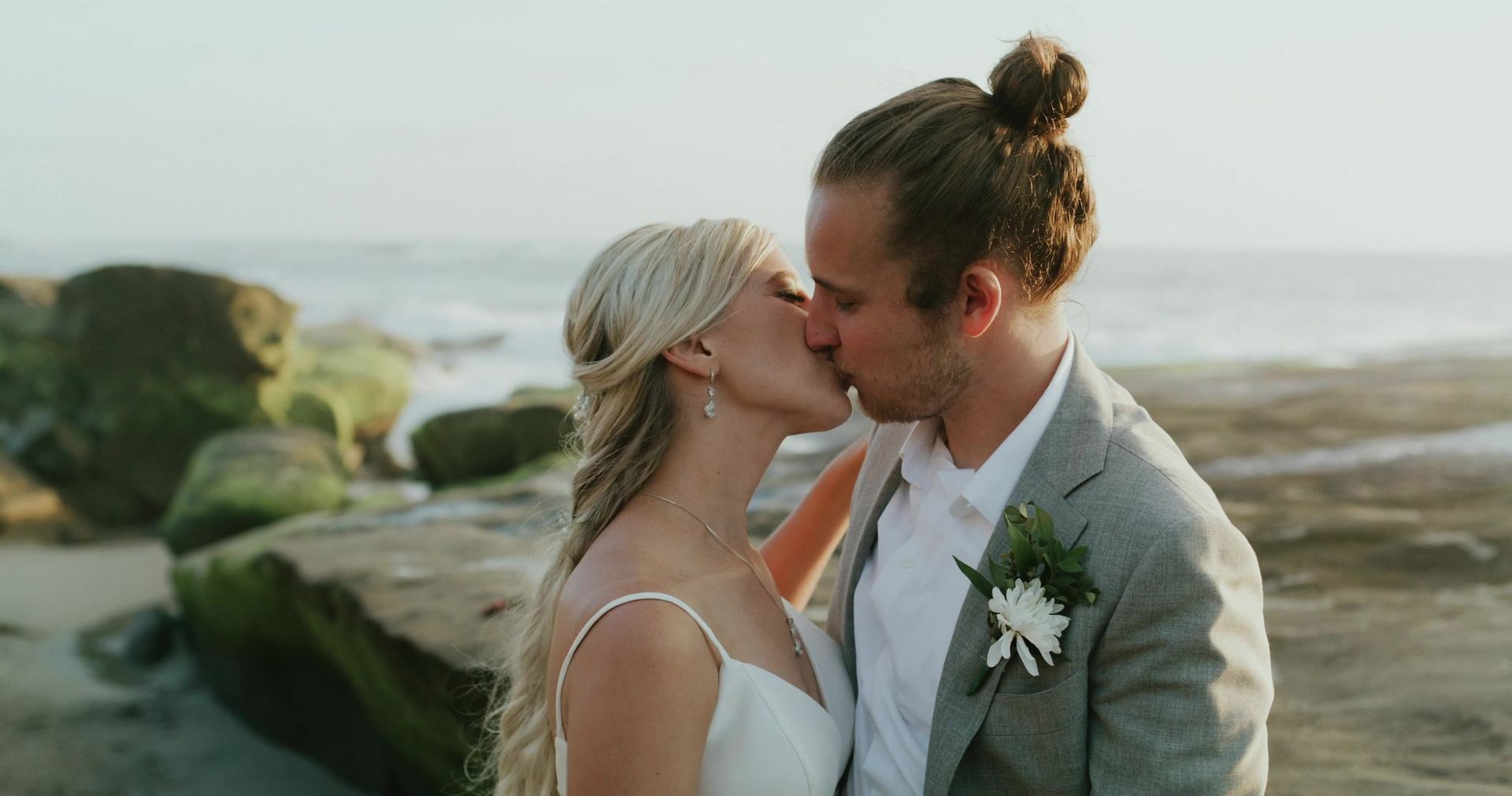 Zoe and Mitch kissing on the rocks at the shoreline, mossy boulders and the water behind them.