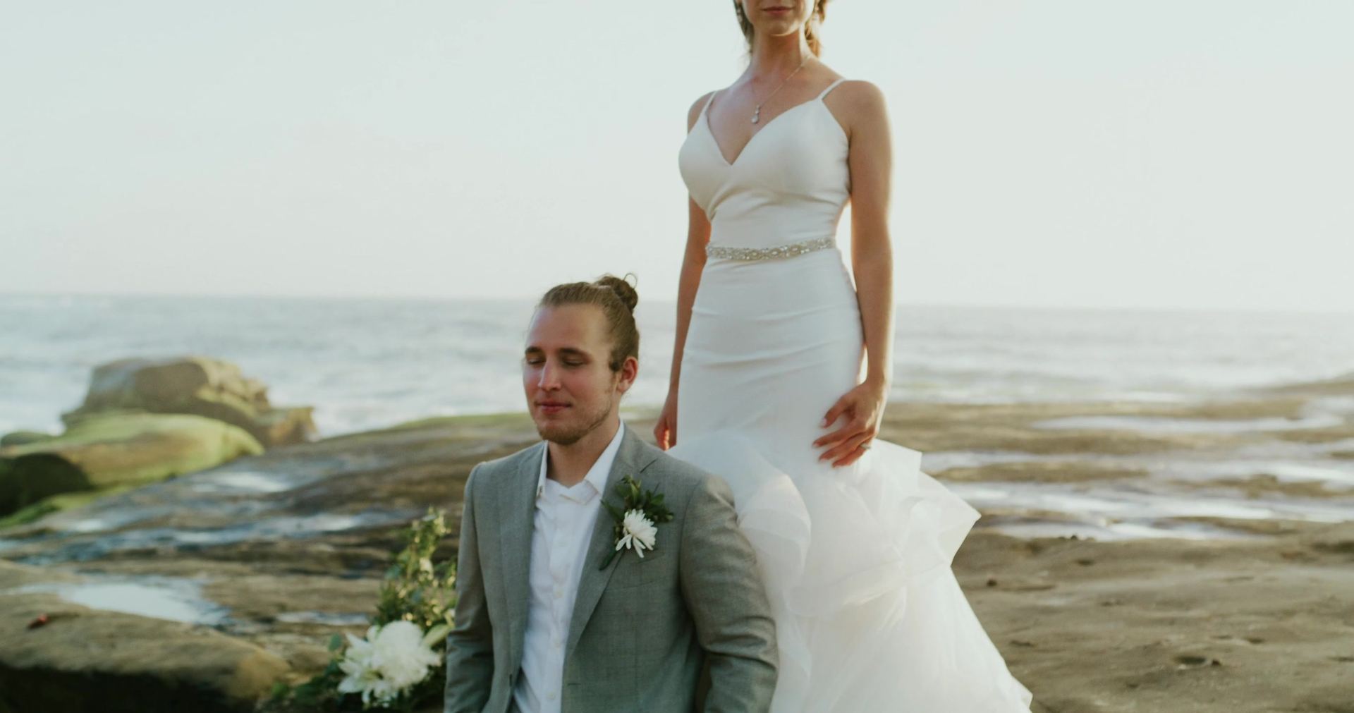 Zoe standing on the rocks above Mitch, who sits in front of her with his eyes closed, the ocean flat behind them.