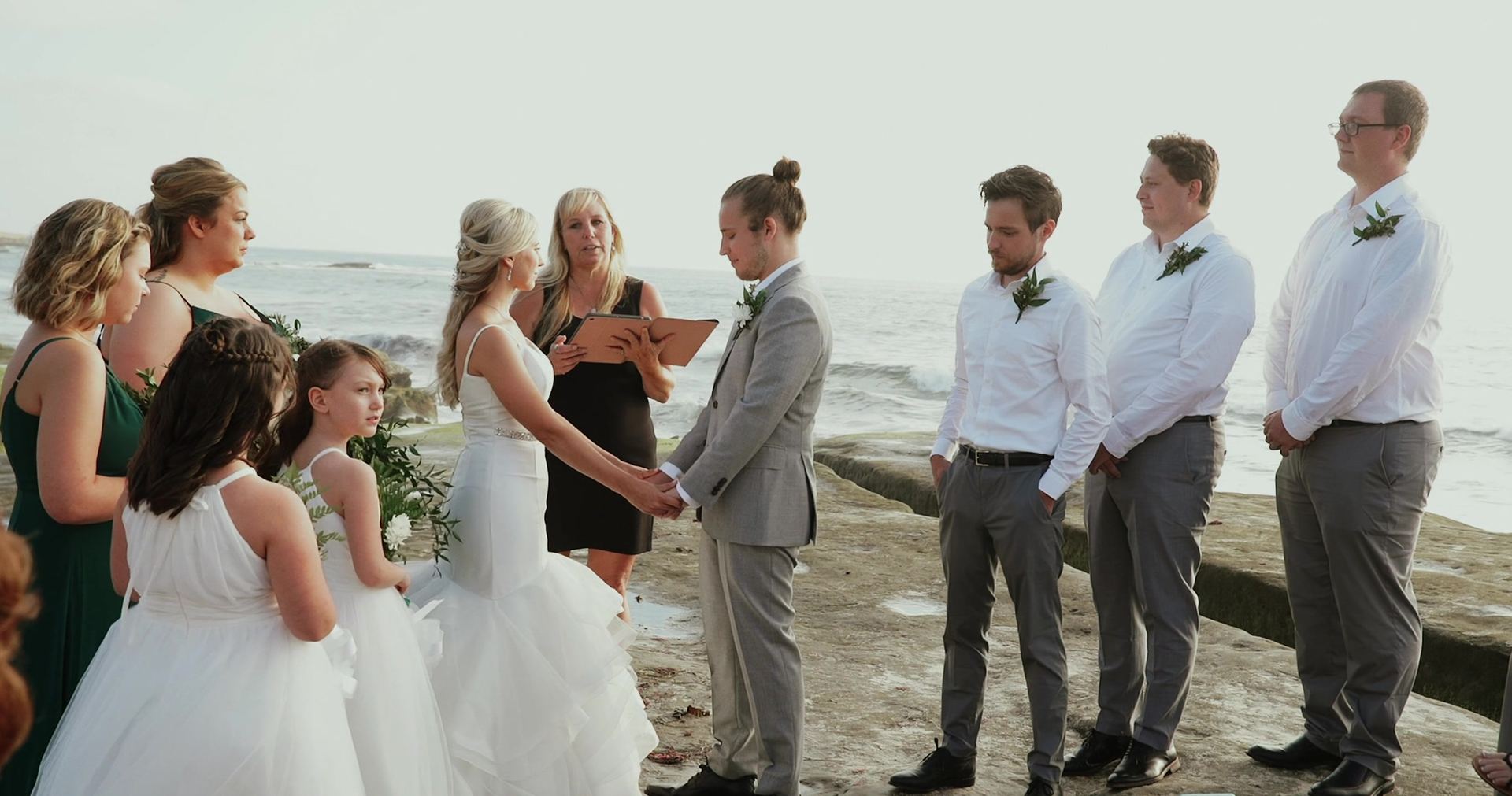 Zoe and Mitch holding hands as the officiant reads from a folder, bridesmaids and two flower girls to one side and the groomsmen to the other.