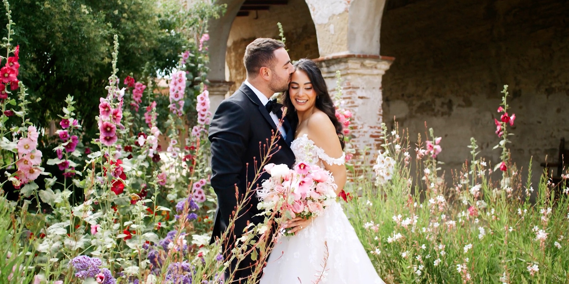 Bride and groom among the tall hollyhocks in the gardens of Mission San Juan Capistrano