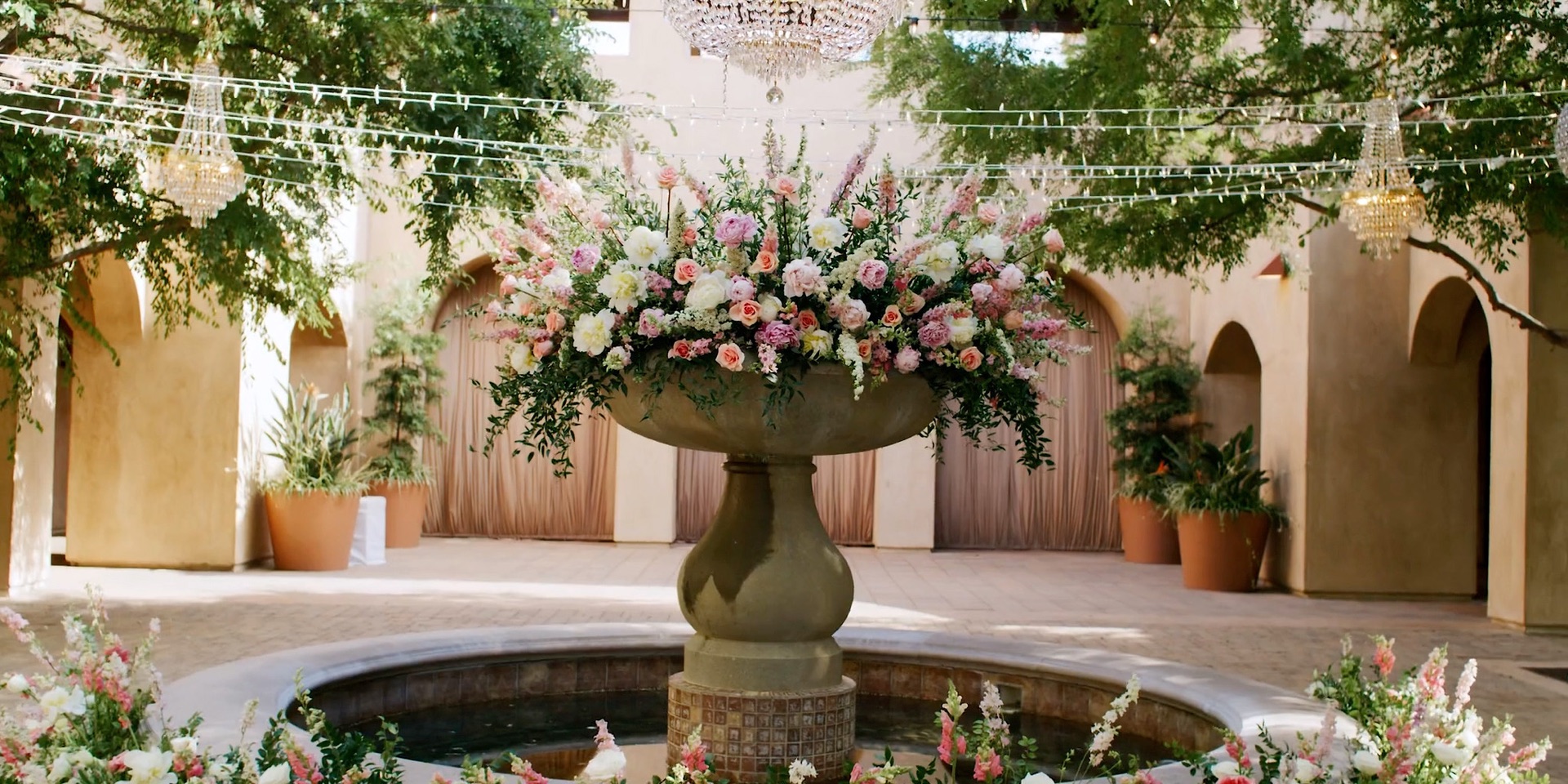 The Serra Plaza courtyard in San Juan Capistrano, its central fountain crowned with roses beneath a canopy of string lights and crystal chandeliers
